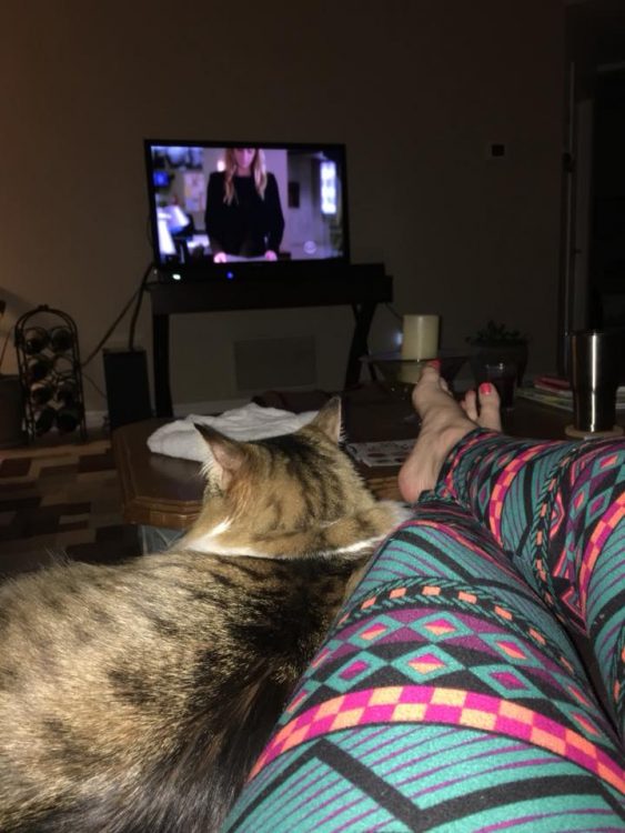 woman lying on her couch in colorful leggings next to her cat and watching tv