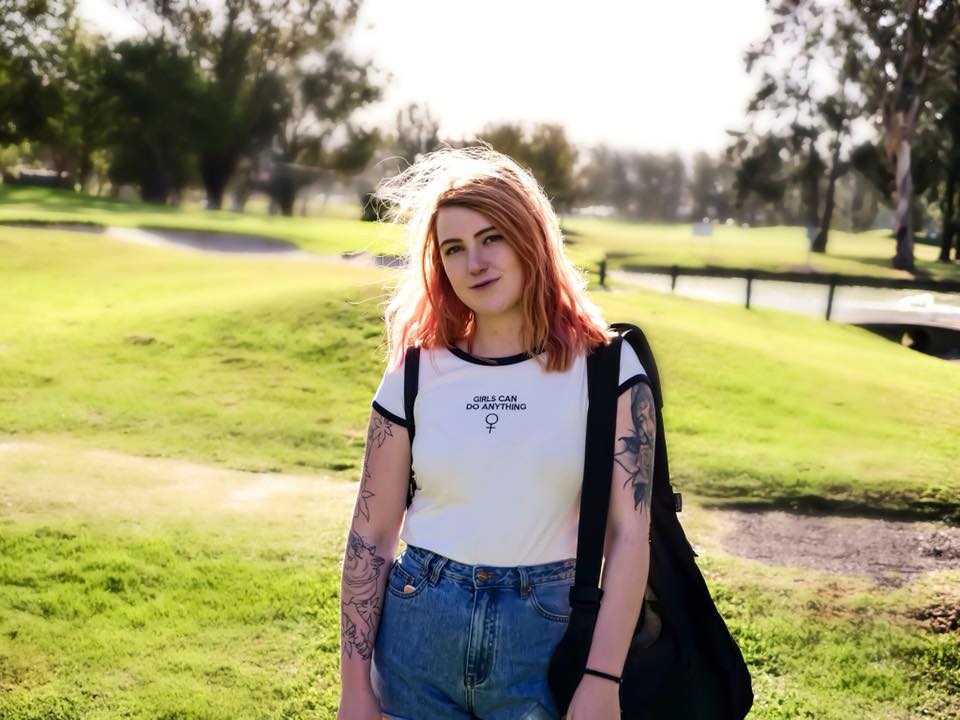 woman with pink shoulder-length hair wearing a shirt that says 'girls can do anything' standing outside in a field holding a black bag