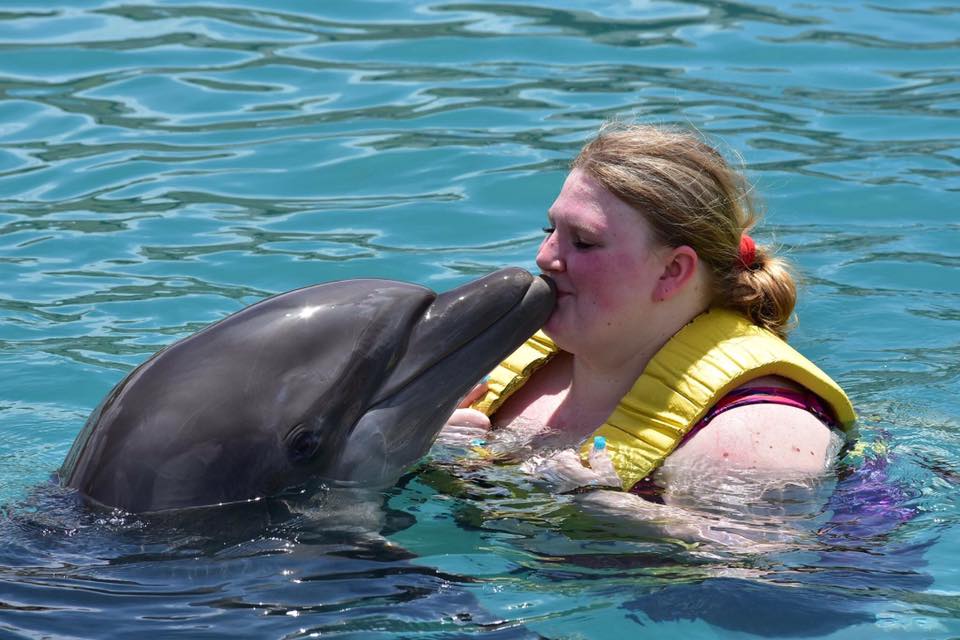 woman in a yellow life jacket kissing a dolphin