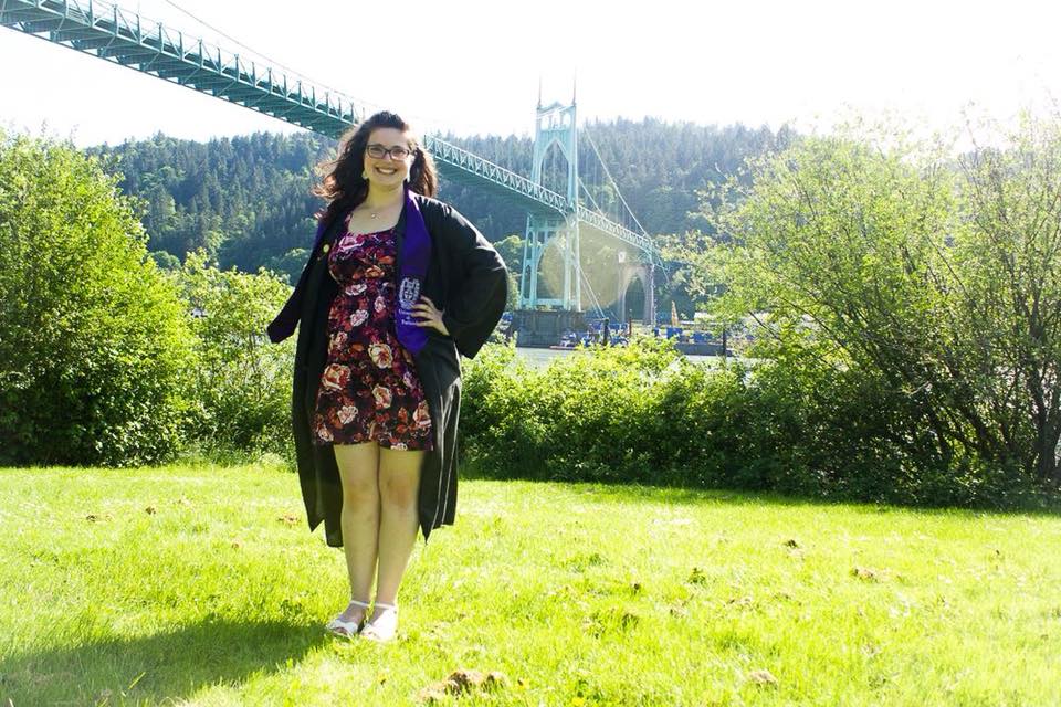 woman wearing her graduation gown and posing in a field with a bridge in the background