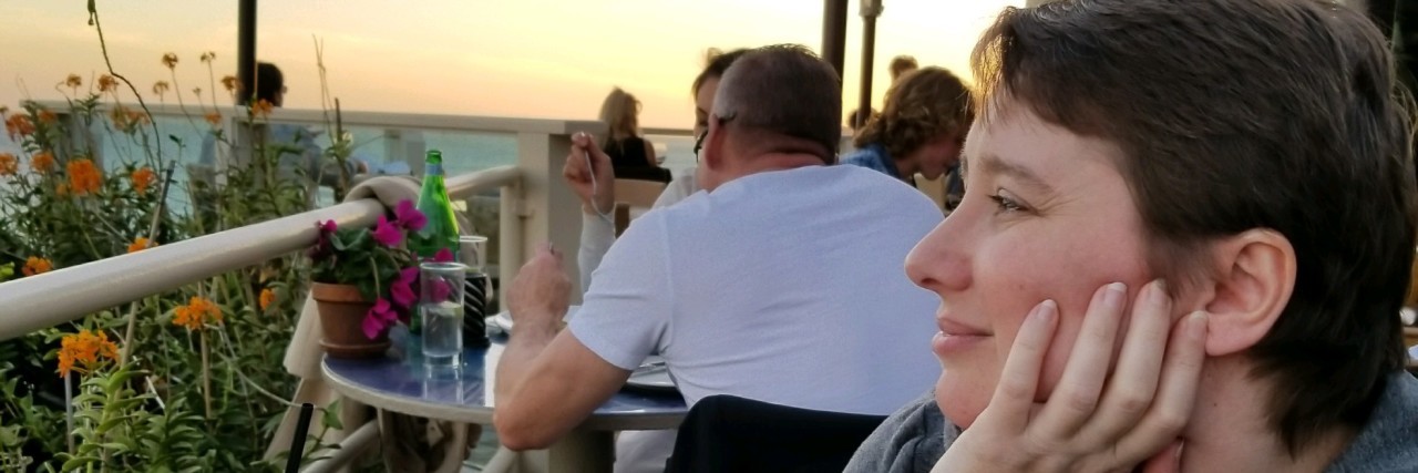Taking Risks and Enjoying Life With Chronic Illness woman sitting on a patio of a restaurant by the ocean looking out at the sunset