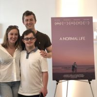 Alex with his brother and mom standing next to movie poster for "A Normal Life."