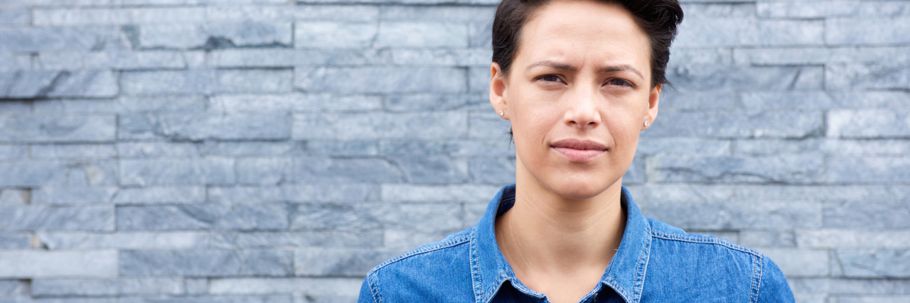 Why You Shouldn't Call Someone With Chronic Illness a Survivor woman with short brown hear wearing a jean jacket and standing in front of a tiled wall looking at the camera