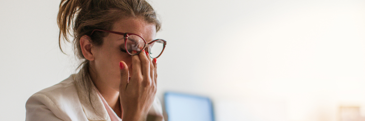 When You're Misunderstood at Work Because of an Illness A woman sitting at her desk, rubbing her eyes.