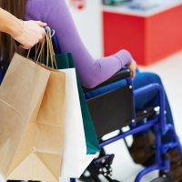 A girl being pushed in a wheelchair, while someone who is pushing her is holding shopping bags.