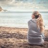 Pensive lonely young woman tourist sitting on beach hugging her knees and looking into the distance with hope