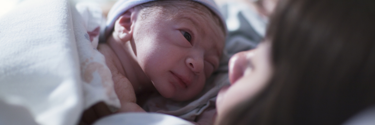 A newborn in a blue cap baby rest on his brunette asian mothers chest and stares into her eyes for the first time.
