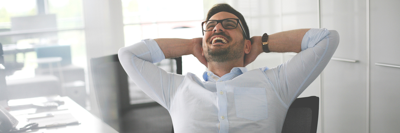 Why You Should Proudly Tell the World You’re ADHD A man sitting at his desk, wish his head back, laughing. He has dark hair, glasses and is wearing a button-up shirt.