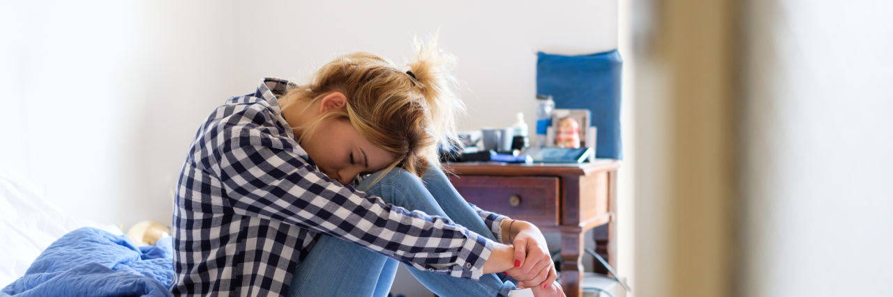 sad young woman sitting on the bed in a messy room with her head in her knees