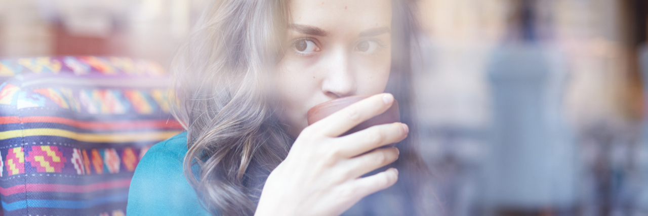 woman with brown hair sitting behind a window sipping coffee