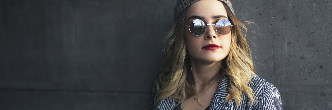 What People Should Know About Adrenal Insufficiency woman standing against a wall wearing a black and white jacket, sunglasses and a hat