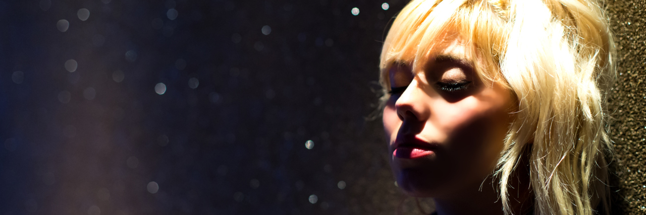 Are You Grieving an Illness? It's OK to Repeat the Stages of Grief. A blonde woman in a dark room with her eyes closed.