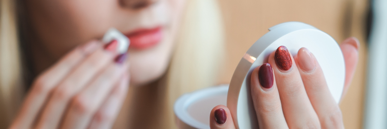 The Ways I’ve Tried to Stop My Compulsive Skin Picking young woman looking at reflection in compact mirror with cotton swab to lips