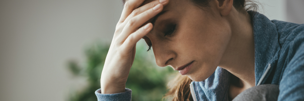 When Others With Your Illness Can Do More Than You woman sitting on her couch at home looking down and resting her head on her hand