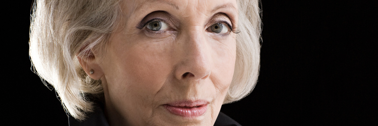 Fibromyalgia: I'm Damned If I Do, and Damned If I Don't An older woman with a serious expression, in front of a black background while wearing a black top.