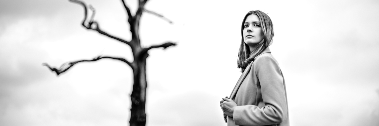 A black and white image of a woman standing alone, near a tree without leaves.