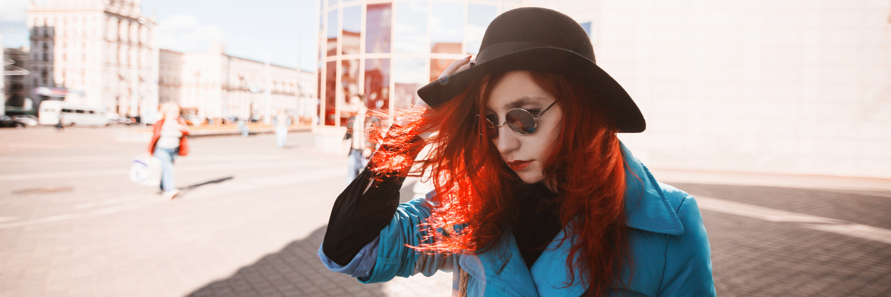 Woman with red curly hair in blue coat and black round sunglasses in city.