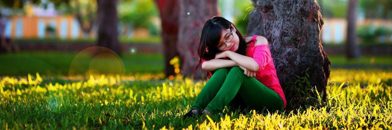 Why It's Hurtful to Call Someone With an Illness 'Weak' woman sitting outside in a field of yellow flowers and leaning against a tree