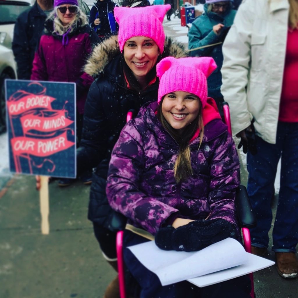 the author sitting in a wheelchair and wearing a pink hat with a woman standing behind her wearing the same pink hat