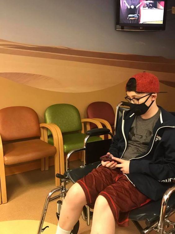 Meg's son, wearing a medical mask and sitting in a wheelchair in the waiting room