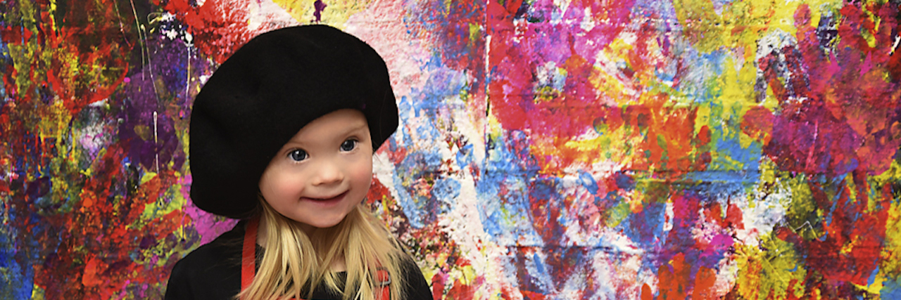 The Importance of Positive Peer Models for Girls With Down Syndrome Girl with Down syndrome dressed as artist wearing red apron and the wall behing is colored in hand prints of different colors, no white spaces.