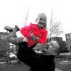 Black and white image of dad lifting daughter ith Down syndrome in the sky, her coat is red, only color in the image