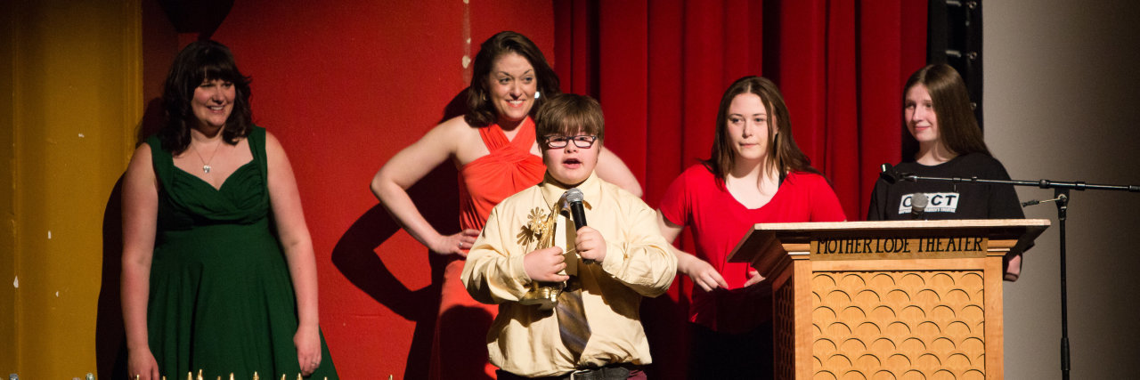 What Community Inclusion Looks Like for My Son With Down Syndrome Boy with Down syndrome speaking into microphone, four teenagers stand behind him, there is a table with trophies