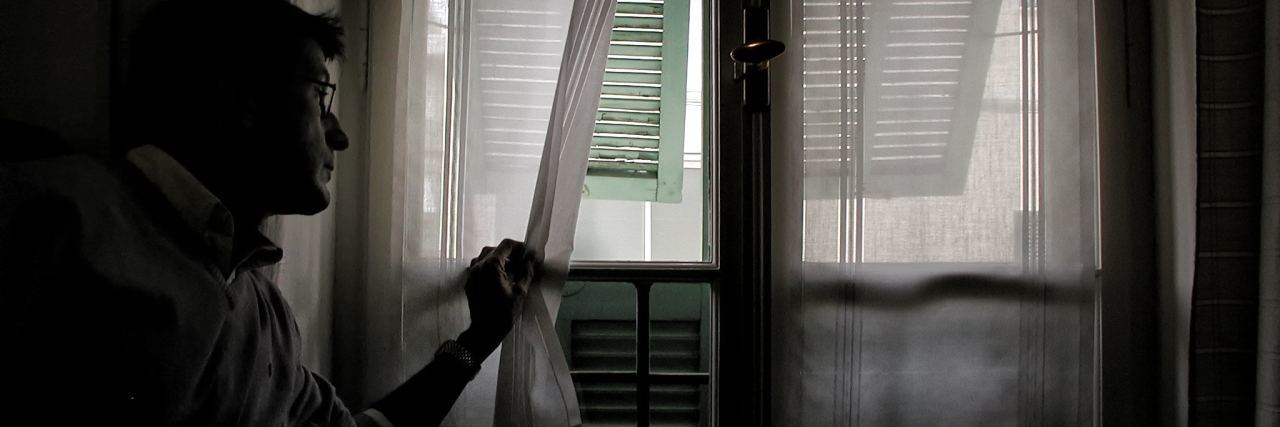 man wearing glasses in dark room and peeking through curtain