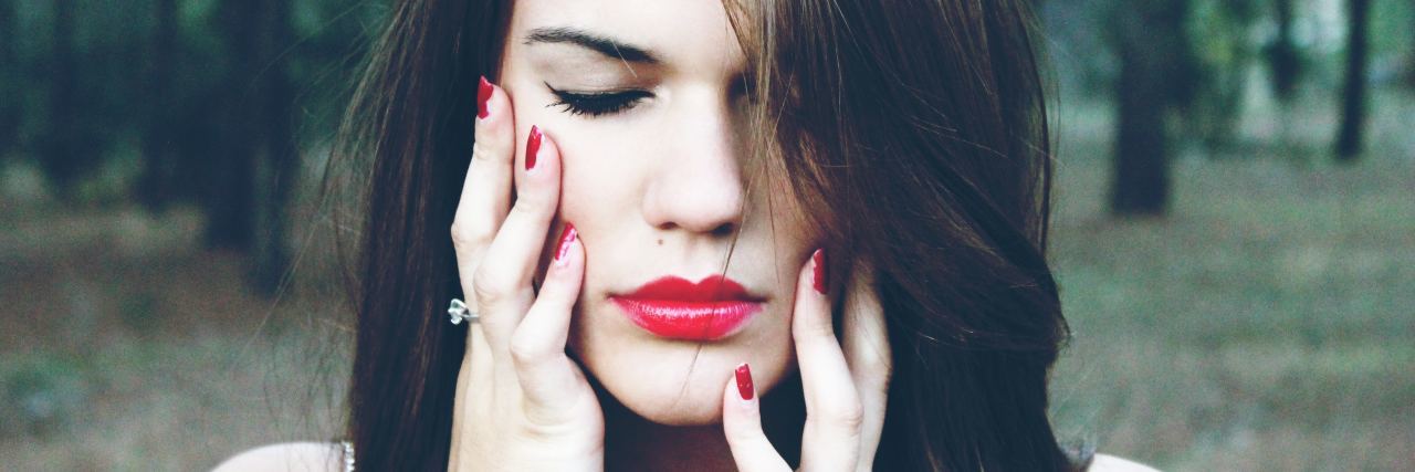 How I Cope With Chronic Pain woman with brown hair wearing red lipstick standing outside in a forest and holding her face