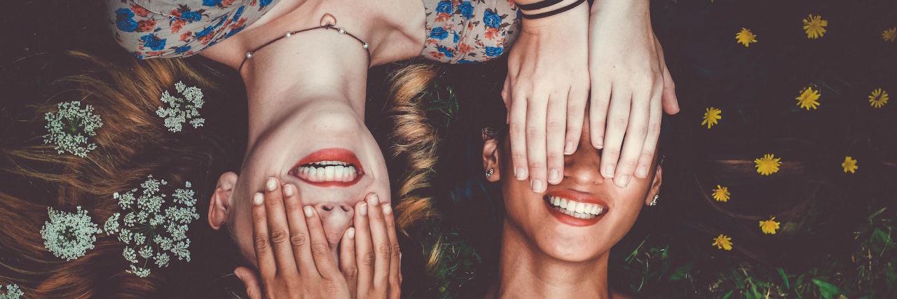 Two grinning women lying on their backs from different directions, their heads close together, and they playfully cover each other's eyes.