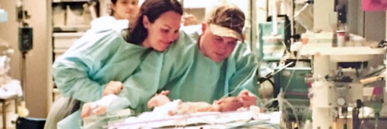 Mother and father leaning over tiny baby at hospital. Parents wear green paper hospital gowns.
