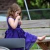 Little girl sitting on park bench, wearing a fancy purple dress, no shoes, and blowing bubbles