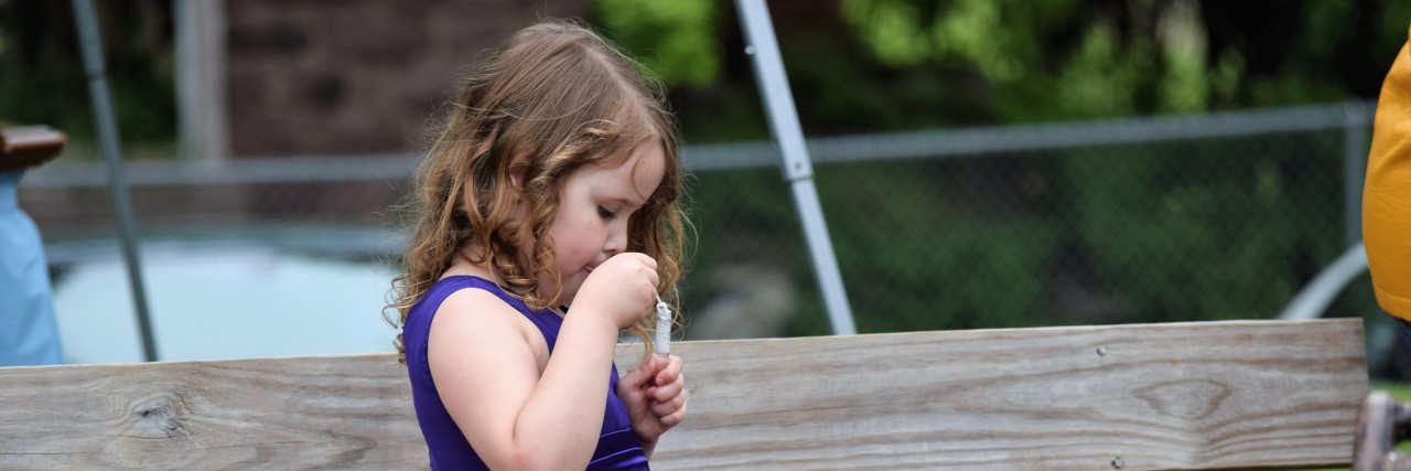 Little girl sitting on park bench, wearing a fancy purple dress, no shoes, and blowing bubbles