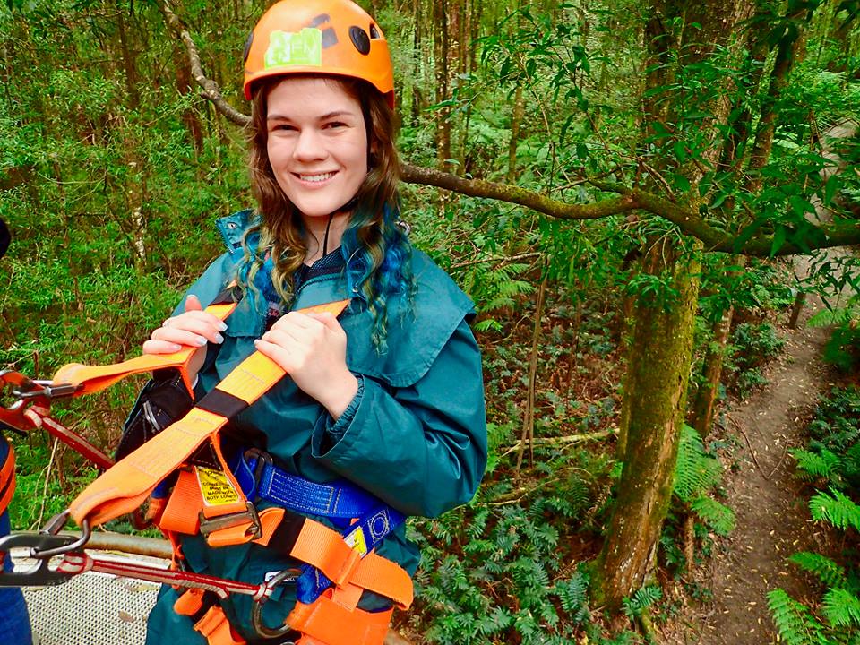 woman doing a zipline course in the trees. she's wearing an orange helmet and turquoise jacket