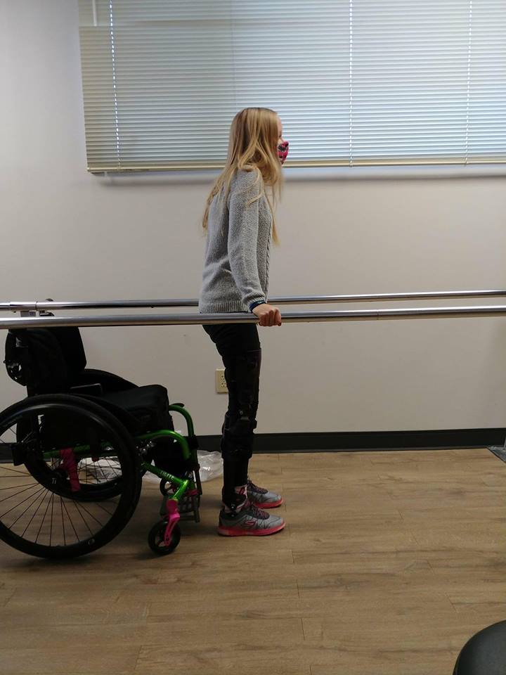 woman learning to walk. she's leaning on two bars with her wheelchair just behind her