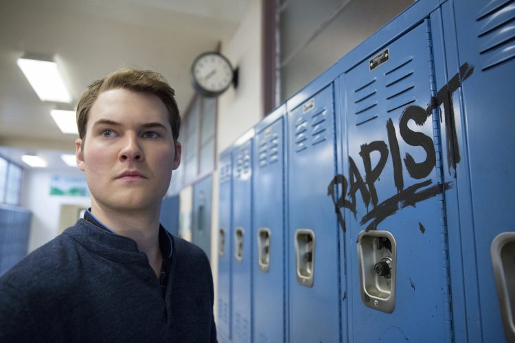Bryce Walker standing in front of his locker. The word "rapist" is grafittied on it.
