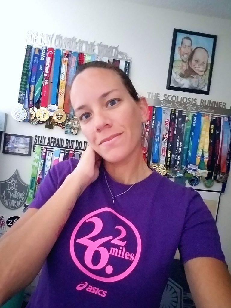 the author wearing her purple 26.2 shirt and standing in front of a wall of medals