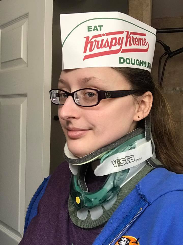 woman wearing glasses, a neck brace and a krispy kreme doughnut hat