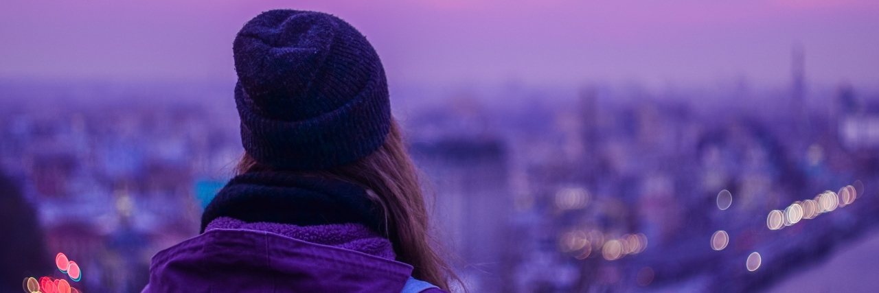 Hipster girl traveler with backpack looking at winter evening cityscape, purple violet sky and blurred city lights
