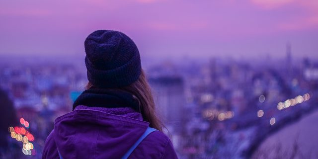 Hipster girl traveler with backpack looking at winter evening cityscape, purple violet sky and blurred city lights