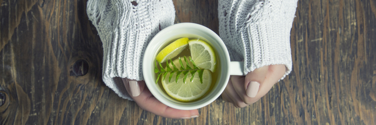 A woman holding a coffee mug, with tea inside it - as well as thyme herb, and lemon slices.