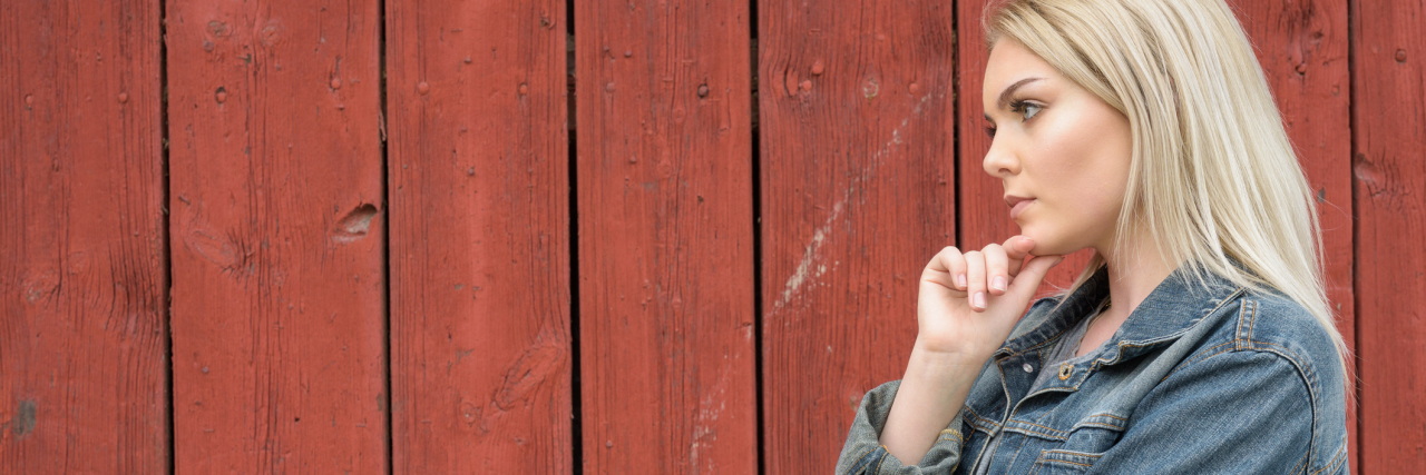 Why I Tell People About My Chronic Illness blonde woman in a jean jacket standing against a red wooden wall