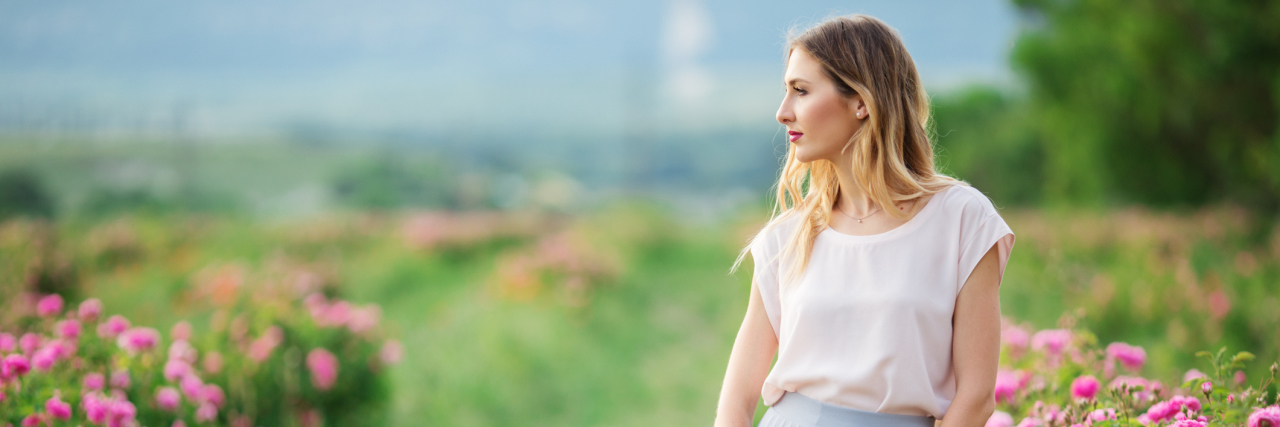 Why I Now Speak Candidly About My Chronic Illness A woman standing outside in a garden, wearing a dress and holding a hat.