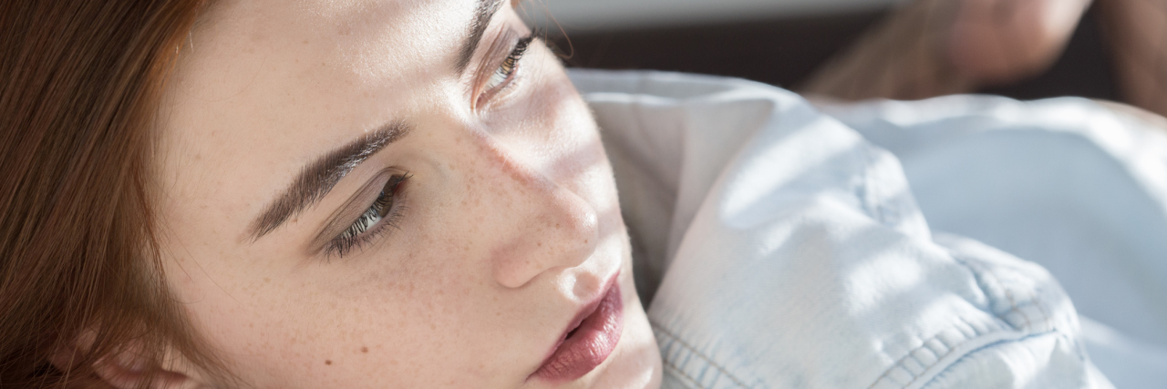 The Truthful Answer I Give When People Ask How I'm Doing A woman laying on her stomach, looking towards a window with the light shining on her.