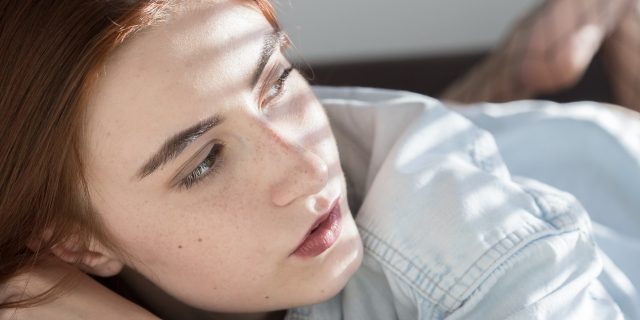 The Truthful Answer I Give When People Ask How I'm Doing A woman laying on her stomach, looking towards a window with the light shining on her.