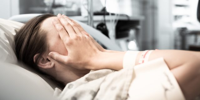 A young woman laying in a hospital bed with her hands to her face.