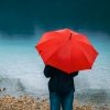 Woman with red umbrella contemplates on rain in front of a lake.
