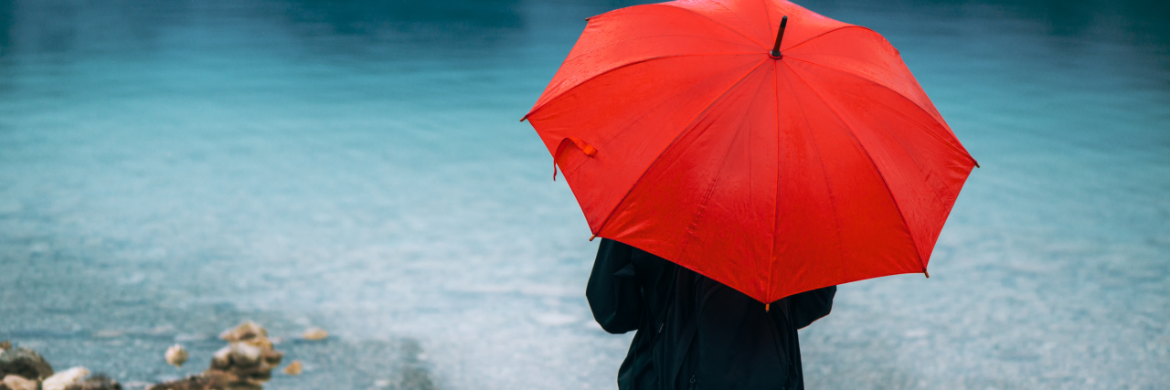 The Lessons You Learn When Living With a Rare Disease Woman with red umbrella contemplates on rain in front of a lake.