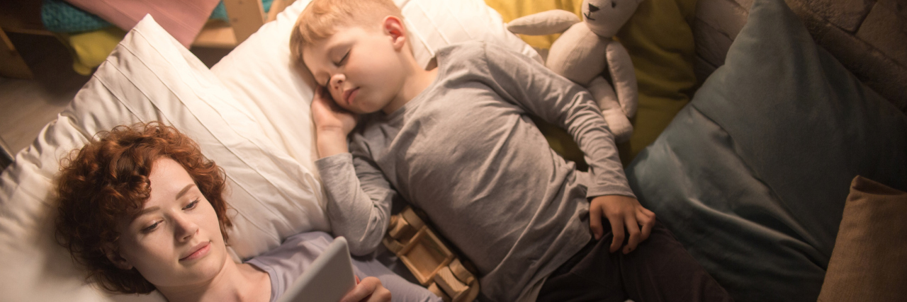 A mom laying in a bed, next to hear young son, while reading on a tablet.