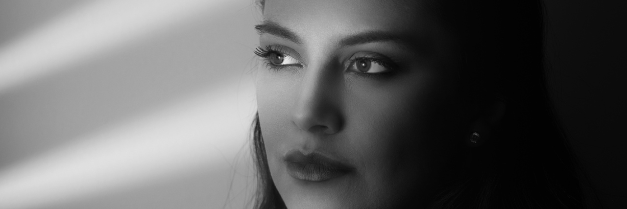 If You're Wondering If Chronic Pain Is 'Really That Bad' A black and white image of a woman looking out a window, only showing the shutter shadows across the wall and across her face.